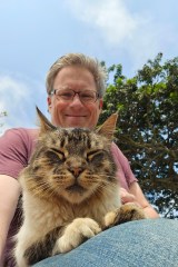 Photo of author Jeff Bogle, sitting with his cat in his lap by a tree. Jeff and the cat are facing the camera, angled down. Jeff is smiling. The cat’s eyes are closed; it looks content.